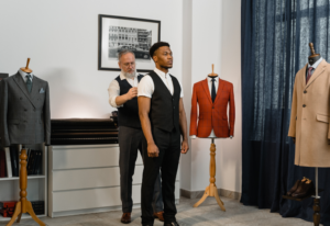 Professional tailor fitting a young man in a black vest at an upscale tailoring shop with suits displayed on mannequins, demonstrating common clothing alterations services.