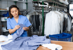 A dry cleaning professional inspects a shirt in a modern facility with clothing on hangers and a rotating conveyor in the background.